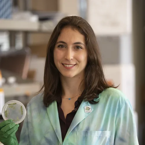 woman in white lab coat, green gloves, holding a petri dish