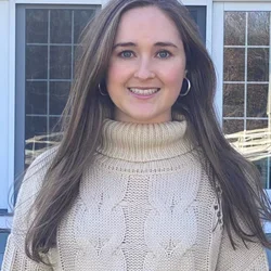 female with brown hair, white sweater, standing in front of a window