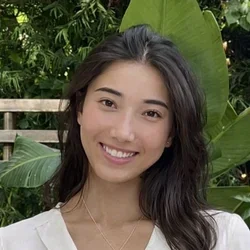female with dark hair wearing a white shirt, standing in front of green plant