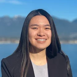 woman in black blazer, grey shirt, standing in front of mountains and water